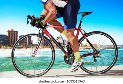 Side View Photo Of A Race Biker, Biking On A Pier In Front Of An Urban Skyline.He Pushes Out Of Th Saddle To Speed Up His Bike.