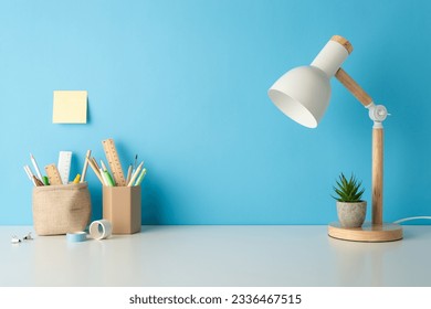 Side view photo of school desk essentials: pen organizers, rulers, lamp, flowerpot, sticky note, and adhesive tape, beautifully set against a blue wall with space for text or advert - Powered by Shutterstock