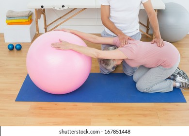 Side view of a physical therapist assisting senior woman with yoga ball in the gym at hospital