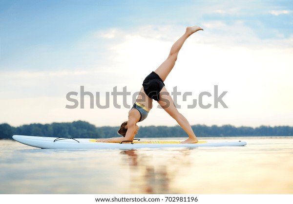 Side view picture of a young\
woman on paddle board practicing three legged down Dog yoga\
pose