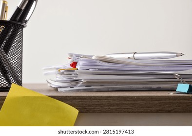 Side view of a pile of office documents with clips and a silver pen resting on top, beside a mesh pen holder on a wooden desk.