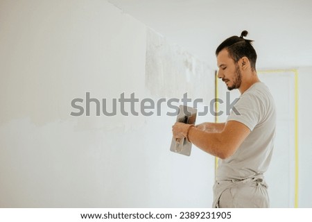 Side view of a plasterer standing in a room in renovation process and using tools for plasterwork and skim coating walls. A worker is renovating house and doing manual building works. Copy space.