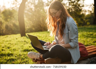 Side view of pleased brunette woman in eyeglasses sitting on bench in park and using laptop computer - Powered by Shutterstock