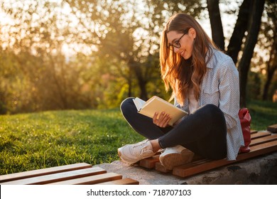 Side view of pleased brunette woman in eyeglasses sitting on bench and reading book in park