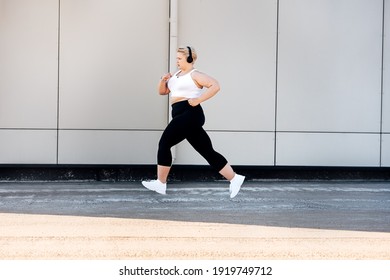 Side view of plus size woman wearing headphones running near a wall. Curvy female in sports clothes jogging outdoors. - Powered by Shutterstock