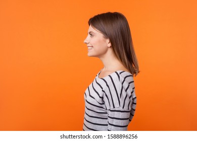 Side View Portrait Of Adorable Happy Young Woman With Brown Hair In Long Sleeve Striped Shirt Standing With Toothy Smile And Looking To The Left. Indoor Studio Shot Isolated On Orange Background