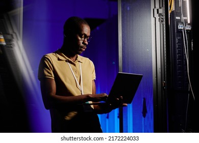 Side view portrait of adult African American man using laptop in dark server room lit by neon light, copy space