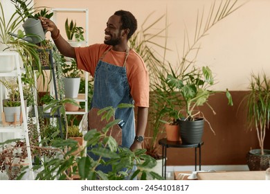 Side view portrait of adult African American man enjoying gardening at home and watering green plants on shelves copy space  - Powered by Shutterstock