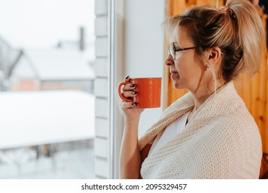 Side View Portrait Of An Adult Pretty Woman Wrapped In Plaid Holding Mug Of Tea Or Coffee And Standing By Window Indoors And Looking At Winter Weather. Home Cozy In Morning In Cold Season Concept.