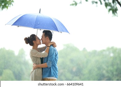 Side View Portrait Of Affectionate Asian Couple Embracing Under Umbrella In Rain, Copy Space