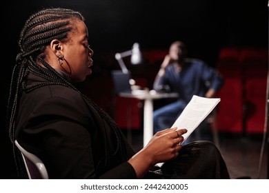 Side view portrait of African American woman reading script while sitting in chair on stage with low lighting copy space - Powered by Shutterstock