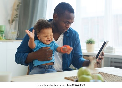 Side View Portrait Of African-American Man Holding Crying Baby While Using Digital Tablet In Home Interior, Copy Space