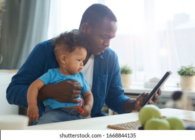 Side View Portrait Of African-American Man Holding Baby While Using Digital Tablet In Home Interior, Copy Space