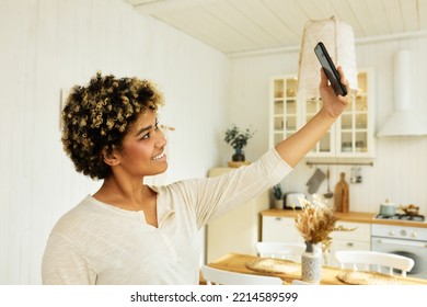 Side View Portrait Of Attractive African American Female Blogger On Influencer Recording Daily Life Stories Or Taking Selfie On Her Front Camera, Holding Phone High Posing In Kitchen Interior