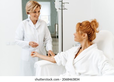 Side view portrait of attractive woman in white bathrobe sitting in armchair with pleasure smile while receiving IV infusion 