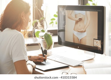 Side view portrait of attractive woman retoucher working on the computer, hand drawing on graphic tablet. Young Caucasian female photographer with red hair in white T-shirt editing photos at home  - Powered by Shutterstock