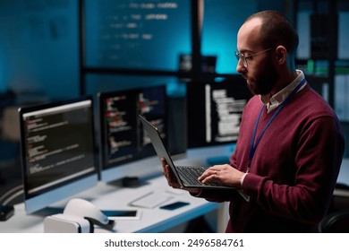 Side view portrait of bearded adult man as computer programmer holding open laptop in IT and cybersecurity office with blue lights copy space - Powered by Shutterstock