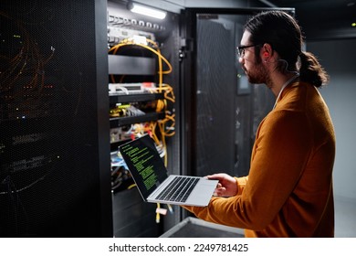 Side view portrait of bearded IT engineer using laptop while setting up network in server room - Powered by Shutterstock - Get 15% off with code: PIKWIZARD15