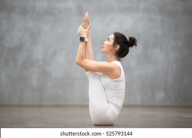 Side view portrait of beautiful young woman working out against grey wall, doing yoga or pilates exercise without mat on wooden floor. Model sitting in upward facing forward bend pose. Full length
