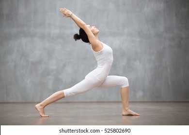 Side view portrait of beautiful young woman wearing white tank top working out against grey wall, doing yoga or pilates exercise. Standing in Warrior one pose, Virabhadrasana. Full length