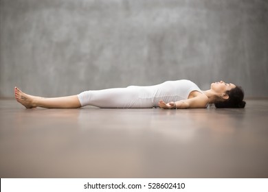 Side View Portrait Of Beautiful Young Woman Working Out Against Grey Wall, Resting After Doing Yoga Exercises, Lying In Savasana (Corpse Or Dead Body Posture), Relaxing. Full Length