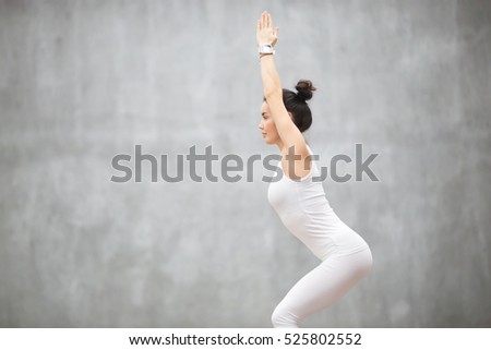 Side view portrait of beautiful young woman wearing white sportswear working out against grey wall, doing yoga or pilates exercise. Standing in Chair, Utkatasana pose. Copy space