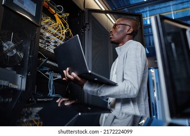 Side View Portrait Of Black Man Working As IT Engineer Holding Laptop While Setting Up Internet Network In Server Room