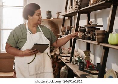 Side view portrait of Black senior woman inspecting pottery pieces on shelf in art studio lit by sunlight copy space - Powered by Shutterstock