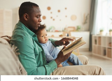 Side View Portrait Of Caring African-American Dad Reading Book To Cute Baby Son At Home, Copy Space