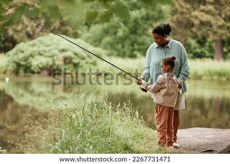 Side view portrait of caring black mother teaching little girl fishing by beautiful forest lake, copy space 