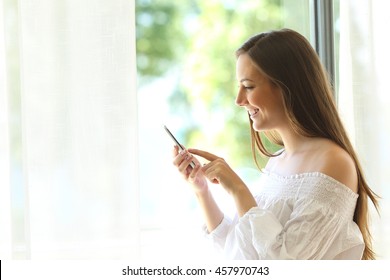 Side View Portrait Of A Casual Girl Using A Smart Phone With A Warm Light Near A Window Of The Living Room At Home