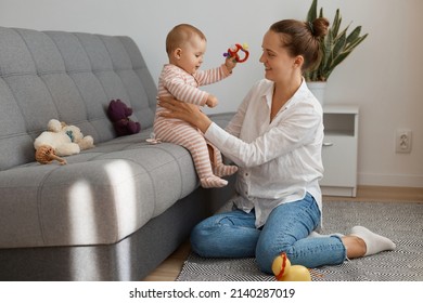 Side View Portrait Of Caucasian Woman Wearing White Shirt And Jeans Sitting On Sofa With Her Baby Daughter, Cute Child Sitting On Cough And Looking With Excitement.