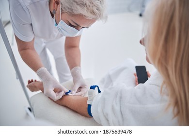 Seitenbild der zertifizierten weiblichen Ärztin, die eine blonde weibliche Patientin auf die intravenöse Vitamin-Therapie in der Schönheitsklinik vorbereitet