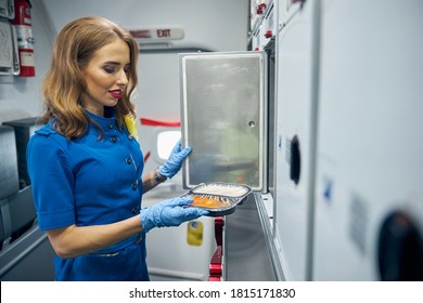 Side View Portrait Of Charming Flight Attendant Standing On The Air Kitchen While Holding Food In Hand