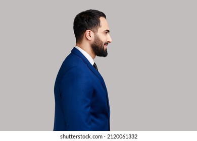 Side view portrait of cheerful bearded man with smile, standing and looking at camera, expressing positive emotions, wearing official style suit. Indoor studio shot isolated on gray background.