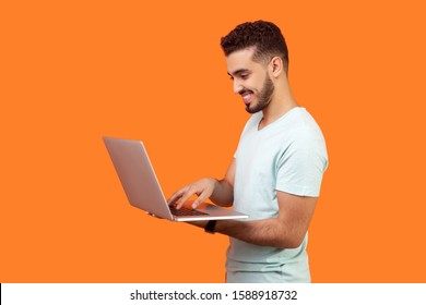 Side View Portrait Of Cheerful Brunette Man With Beard In White T-shirt Working On Laptop Typing Email Or Chatting In Social Network, Happy Face Expression. Studio Shot Isolated On Orange Background