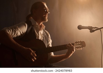 Side view portrait of classic mature guitarist playing acoustic guitar during stage performance in soft spotlight, copy space - Powered by Shutterstock