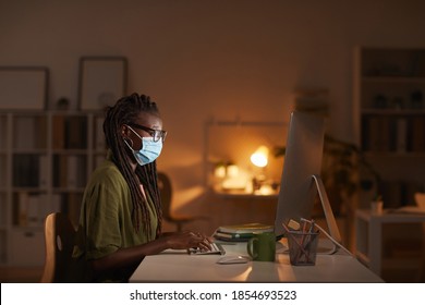 Side view portrait of contemporary African-American woman wearing mask while working in office late at night lit by computer screen, copy space - Powered by Shutterstock