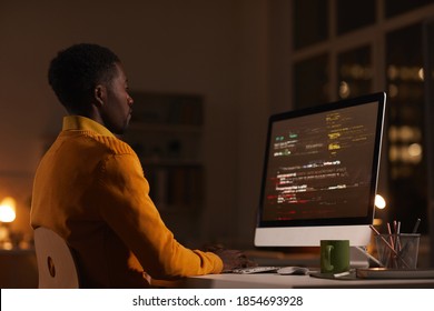 Side view portrait of contemporary African-American man looking at computer screen while working late at night writing code, copy space