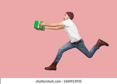Side view of portrait of crazy bearded young hipster man in white shirt and casual hat jumping, running and hurry up with delivery green present. Indoor, isolated, studio shot, pink background