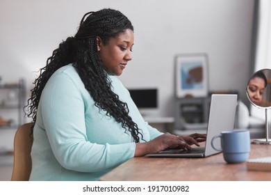 Side view portrait of curvy African American woman using laptop at desk and typing while enjoying work from home in minimal interior, copy space