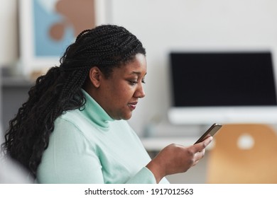 Side view portrait of curvy African American woman looking on smartphone screen while browsing internet sitting on couch in minimal home interior, copy space