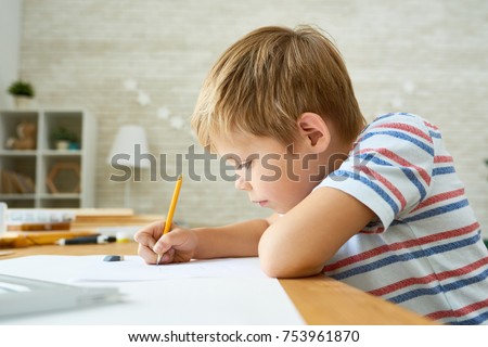 Side view portrait of diligent little boy writing or drawing carefully sitting at desk and doing homework, copy space