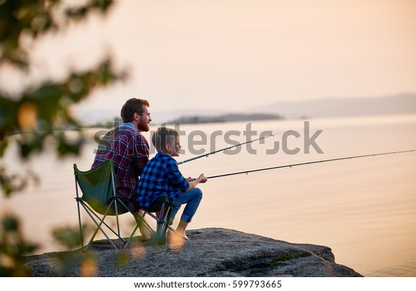 Side view\
portrait of father and son sitting together on rocks fishing with\
rods in calm lake waters with landscape of setting sun, both\
wearing checkered shirts, shot from behind\
tree