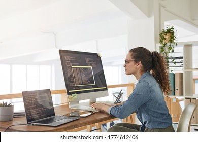 Side view portrait of female IT developer typing on keyboard with black and orange programming code on computer screen and laptop in contemporary office interior, copy space