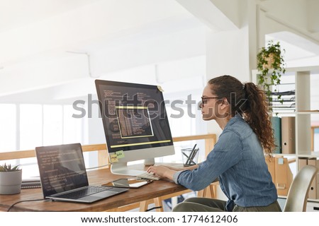 Side view portrait of female IT developer typing on keyboard with black and orange programming code on computer screen and laptop in contemporary office interior, copy space