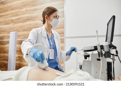 Side view portrait of female doctor using ultrasound machine and wearing mask while examining pregnant woman in clinic