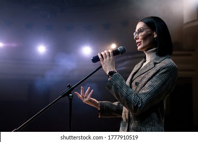 Side View Portrait Of A Female Public Speaker Speaking At The Microphone. Female Entertainer, Presenter Or Actor On Stage. Smoke On Background Of Spotlight. 