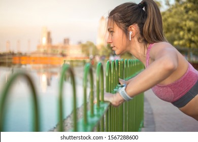 Side view portrait of fitness woman in sportswear with airpods doing push up over fence outdoors