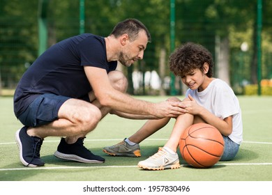 Side View Portrait Of Focused Adult Man Coach Helping Boy With Knee Trauma After Playing Basketball On The Court, Sad Player Feeling Pain, Touching Leg. Activity And Sports Injury Concept.